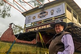 A man in a cowboy hat stands in front of a 100 year old boat surrounded by trees.