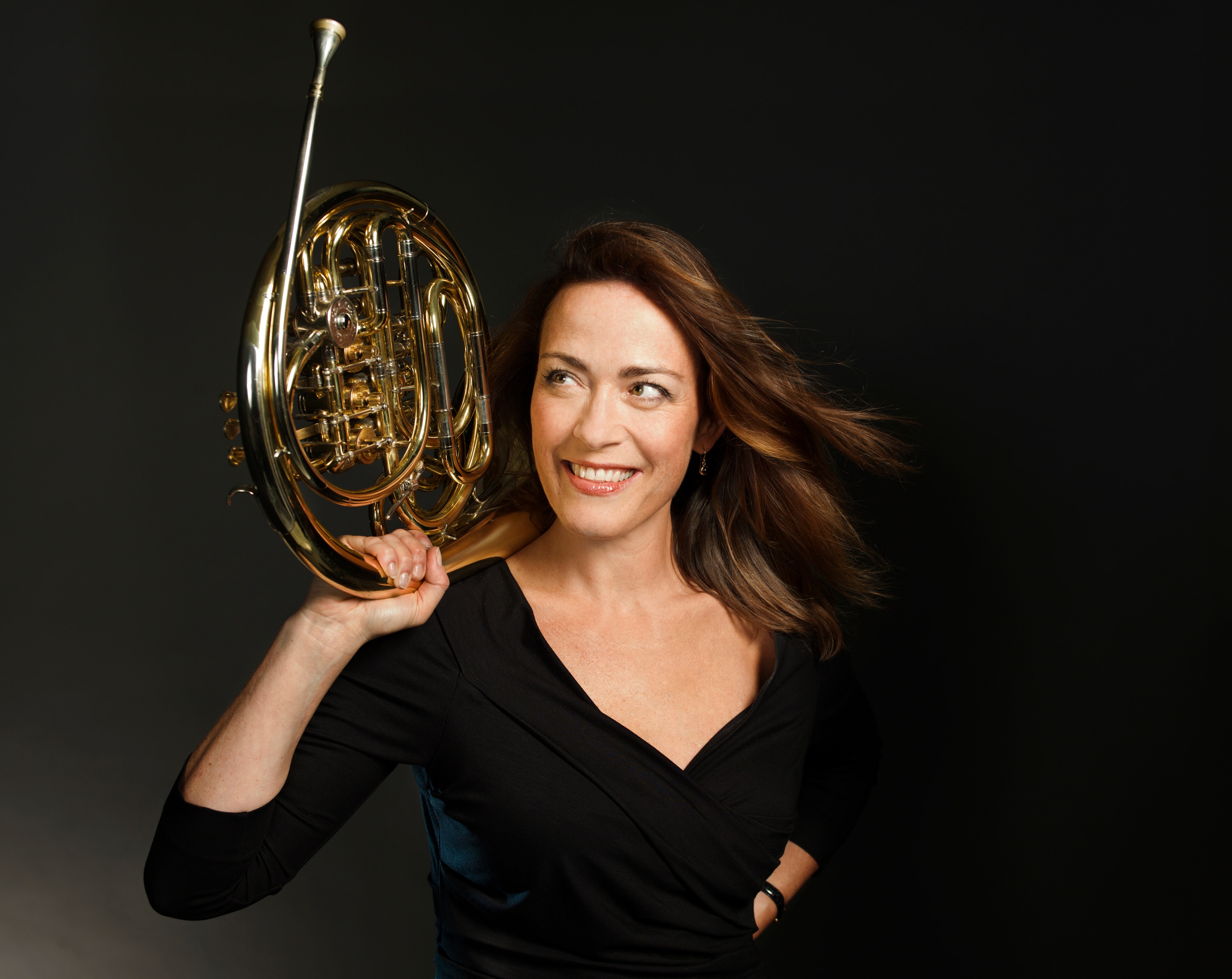 Sarah Willis rests a gold horn on her right shoulder and looks upwards. She wears black against a black background.
