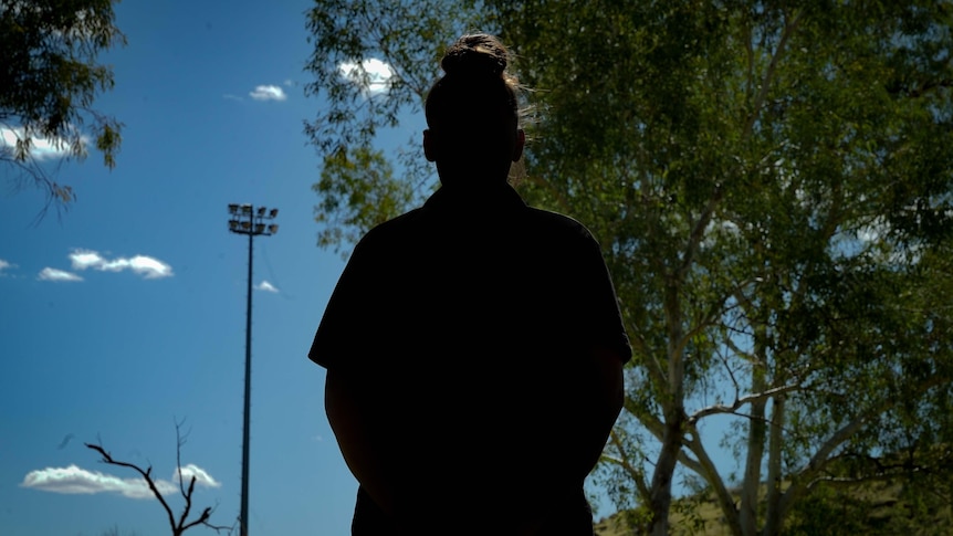 A silhouette of a woman. Her hair is in a bun and she is outside, looking out onto bush.