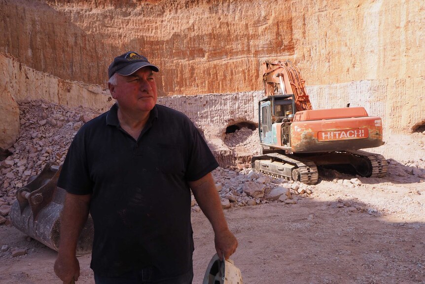 A man stands holding a pick and hard hat in an open-cut mine, with an excavator in the background