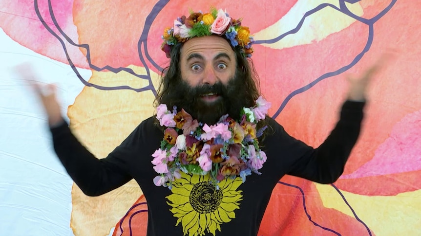 Gardening Australia's Costa with flowers in his hair and beard in front in a pink artsy wall