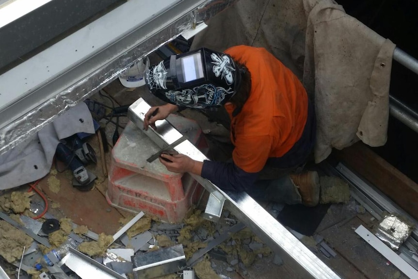 A man in a welding mask surrounded by grey particles from broken roof panels.