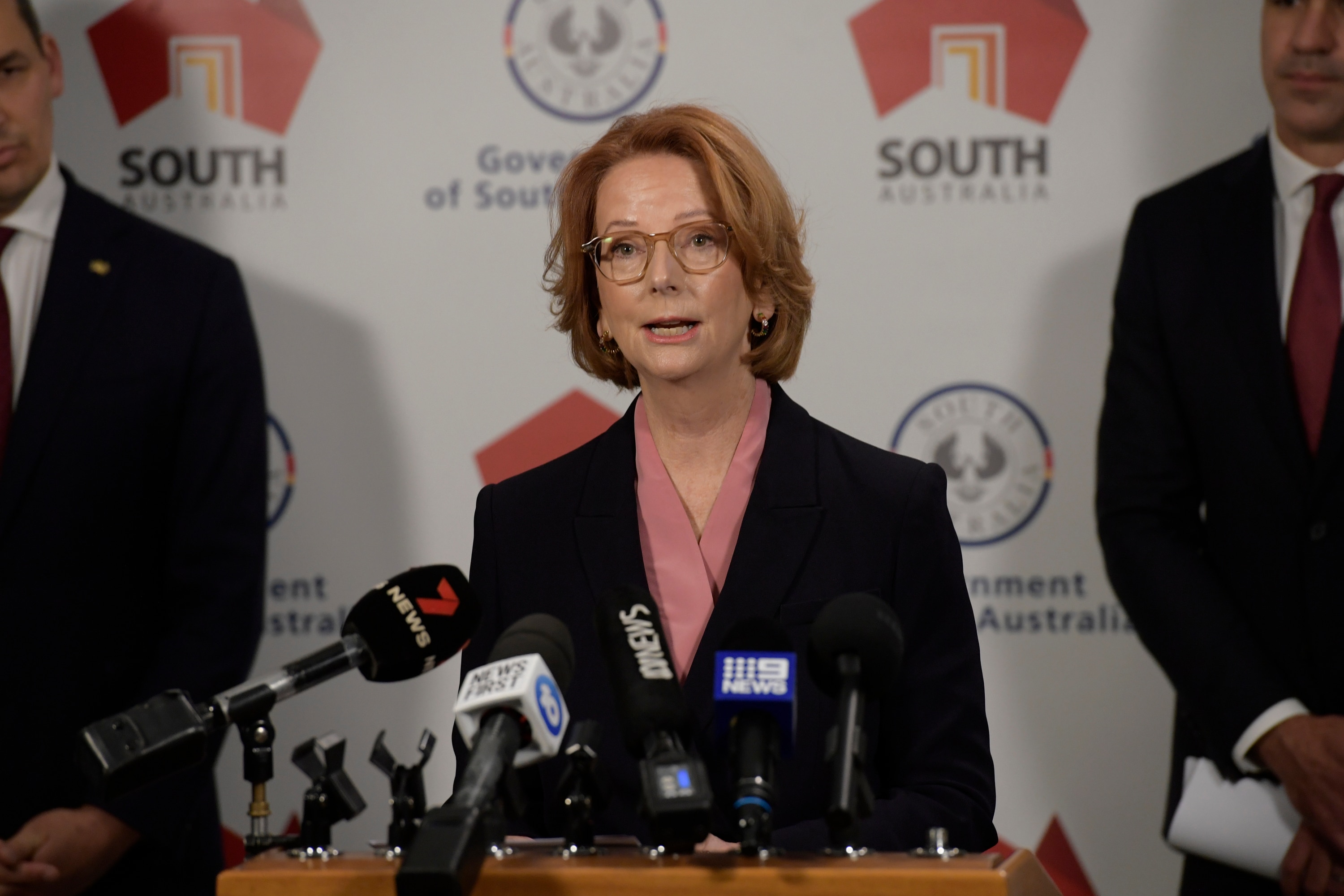 Julia Gillard, a red-headed woman in a suit, speaks at microphones during a press conference.