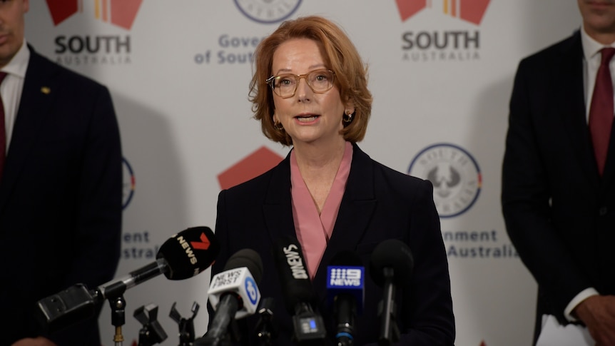 Julia Gillard, a red-headed woman in a suit, speaks at microphones during a press conference.