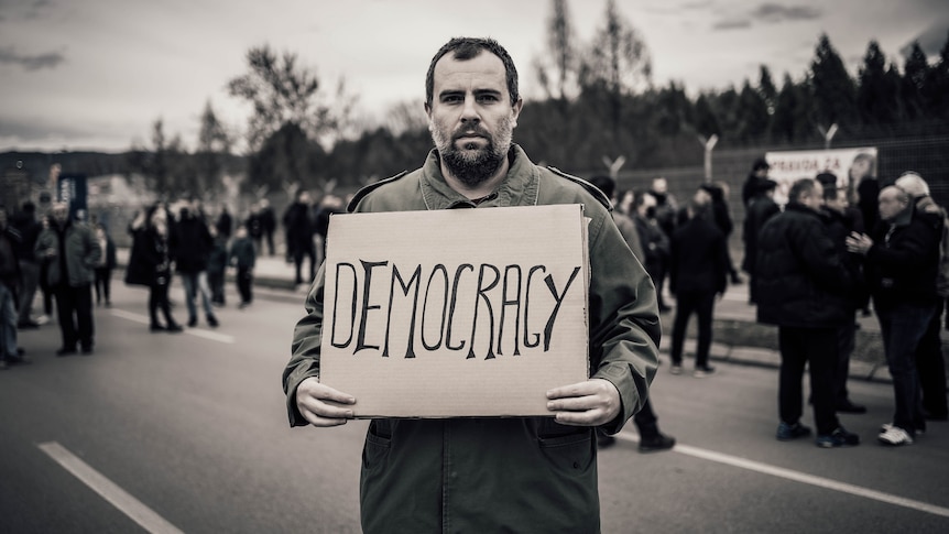 Male protester holding placard with text "Democracy" on the city street. Black and white.