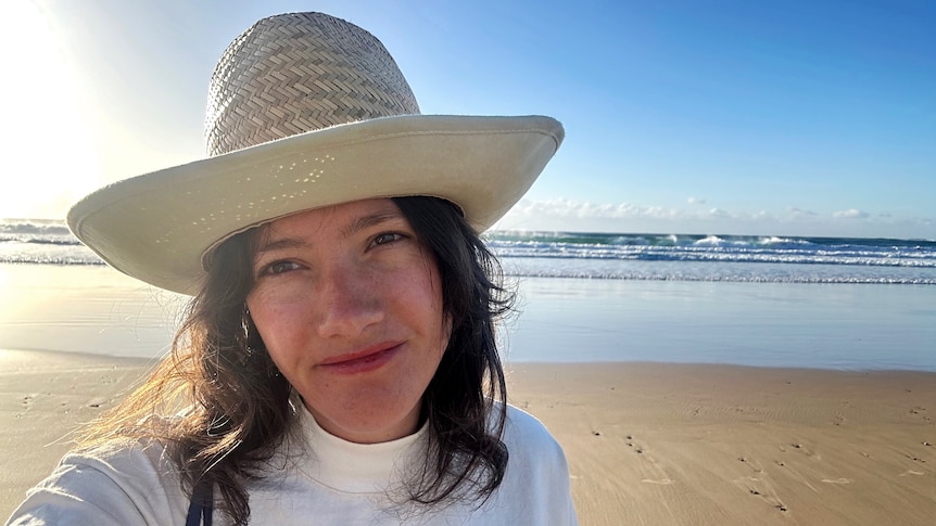 Wearing a straw beach hat and white rashy, Mel Fulton stands on Sawtell beach with the waves behind her