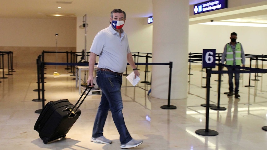 US Senator Ted Cruz, wearing a Texas flag face mask, carrying luggage through an airport.