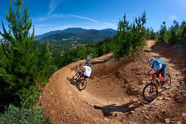 mountain bikers descend on a sharp turn with mountains in the distance. 