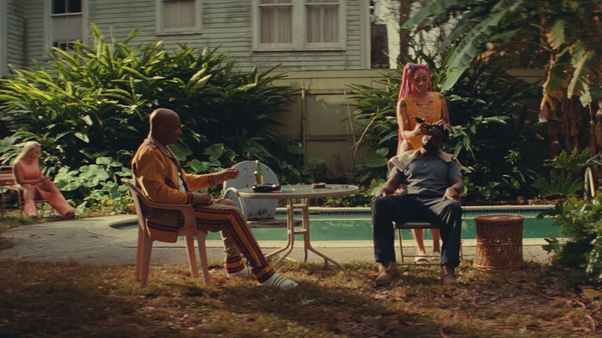 Outdoor shot of two men and two women in a backyard, one sitting on lawn chair having hair clippered while talking to the other.