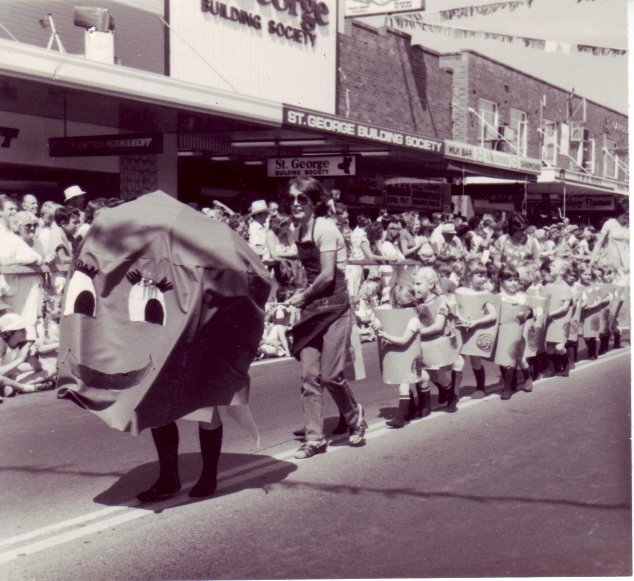 A black and white photo of a giant orange costume walking in a parade, following by children.