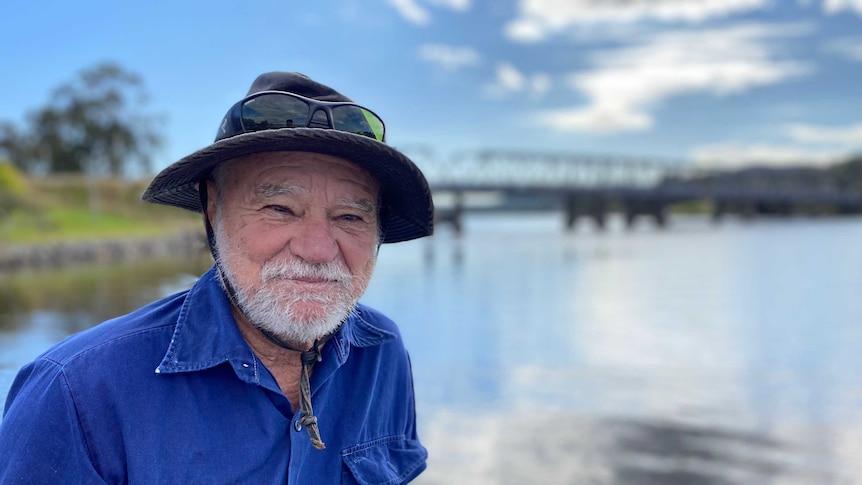 A man stands in front of a river with a bridge in the background.