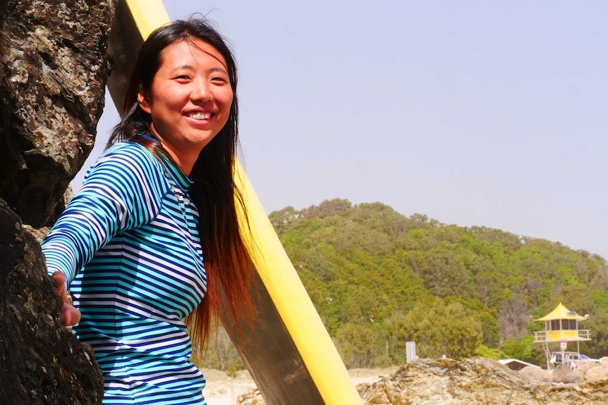 A smiling woman in a stripy top stands next to a surfboard at the beach.