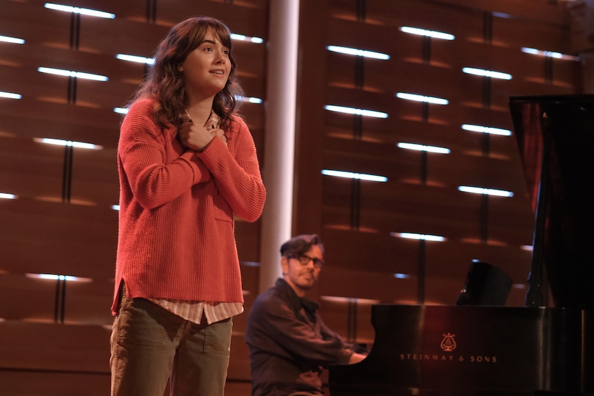 A teenage girl stands on stage smiling, her hands clutched to her chest. A man sits behind her at a piano.