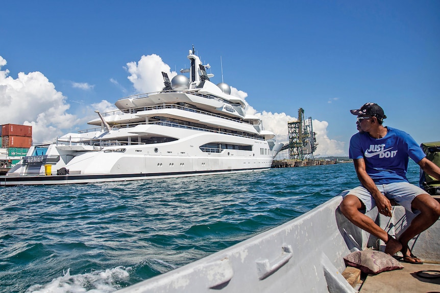Un hombre con camisa azul, sudadera con capucha y gafas de sol se sienta junto al agua con un gran barco al fondo.
