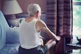 The back view of an elderly man sitting on a couch with his bands on his knees looking out a window onto greenery.