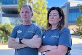 A man and woman wearing blue tshirts stand with their arms folded in front of a hospital