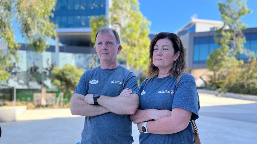 A man and woman wearing blue tshirts stand with their arms folded in front of a hospital