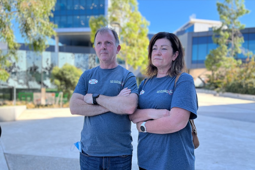 A man and woman wearing blue tshirts stand with their arms folded in front of a hospital