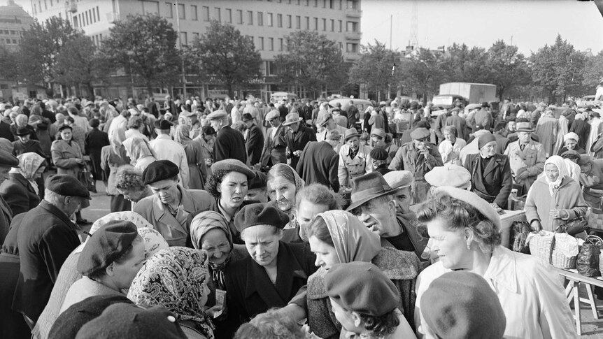 A crowd of middle-aged and elderly Finnish women gather in a circle bartering for an object.