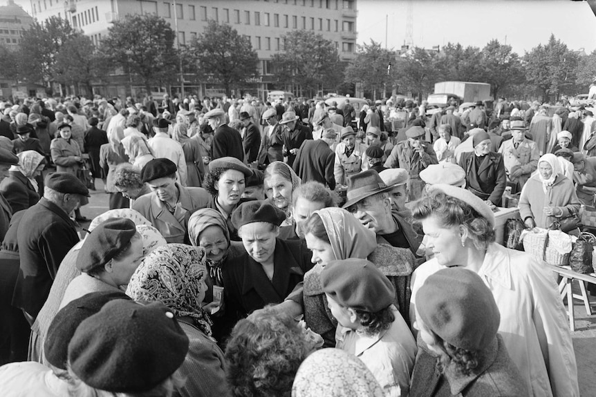 A crowd of middle-aged and elderly Finnish women gather in a circle bartering for an object.