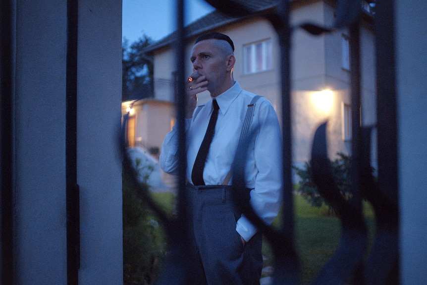 A film still of Christian Friedel, dressed in a 40s-style suit, smoking outside. He is seen through a fence.