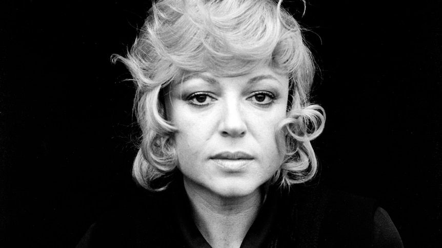 A black and white portrait shows a woman dressed in black with a neutral expression from the shoulders up facing the camera