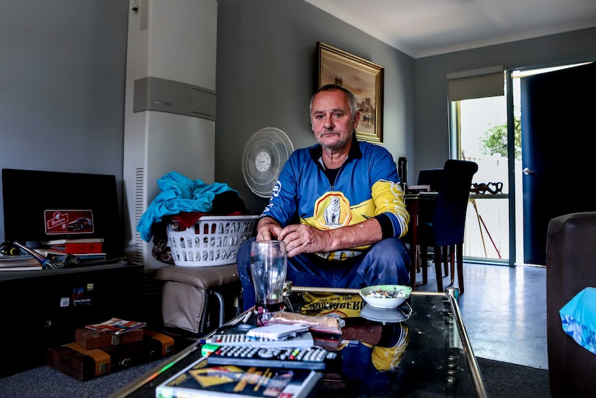 Jamie Cox wearing blue and yellow shirt sits in loungeroom with glass table in front and chair and tables behind