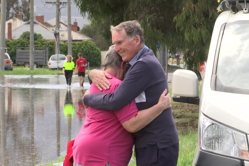 A man and woman embrace, with floodwaters in the background.