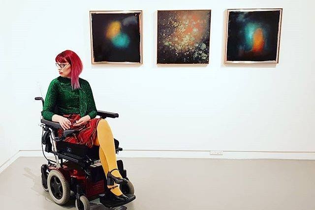 Michelle Roger in a wheelchair at an art gallery. She wears yellow tights, tartan red skirt, green glittery long sleeved top.