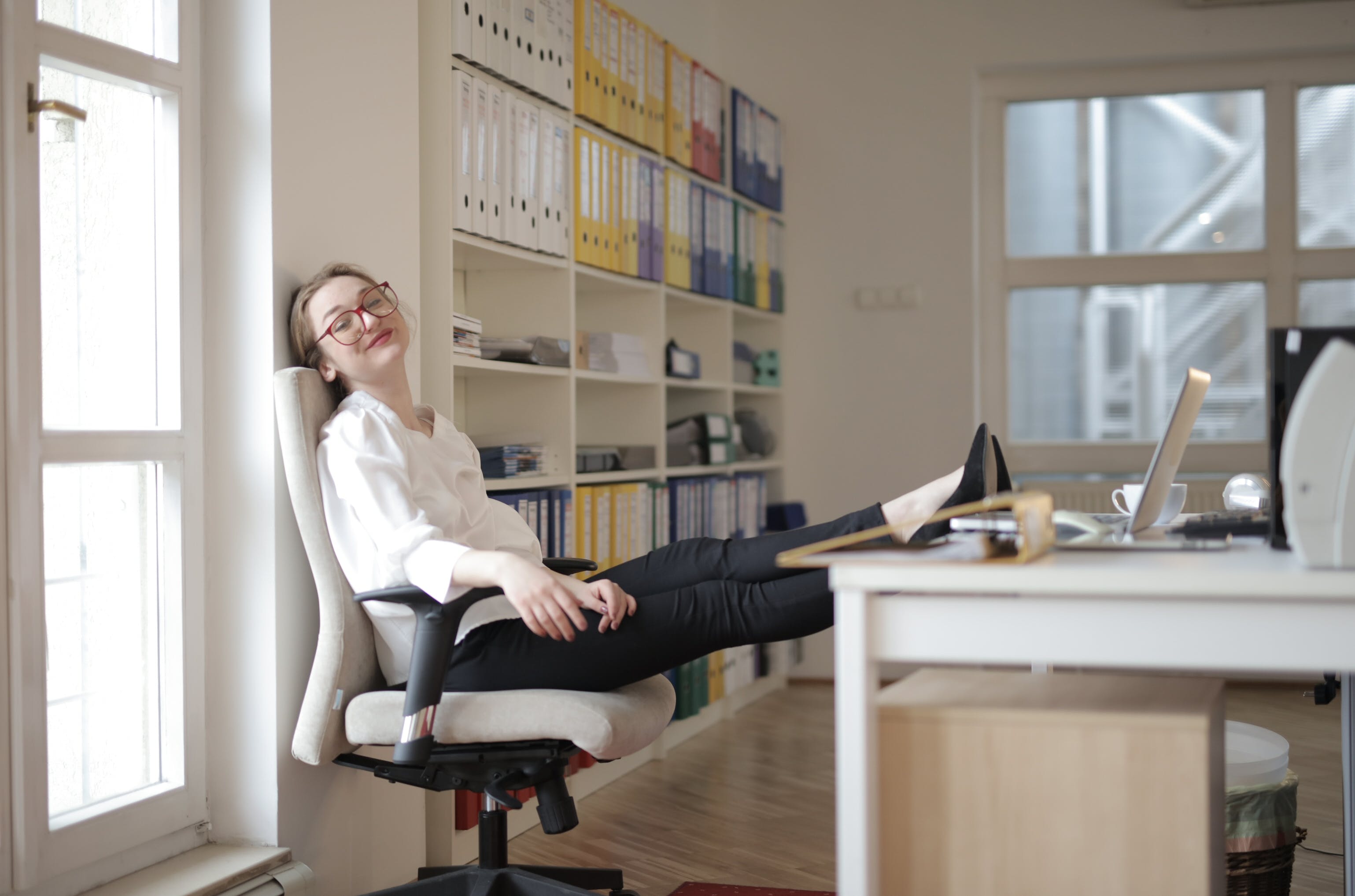 A woman wearing white and black work wear and red glasses reclines on an office chair in a bright office, her feet on the desk.