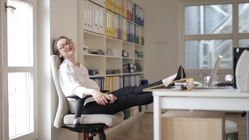 A woman wearing white and black work wear and red glasses reclines on an office chair in a bright office, her feet on the desk.
