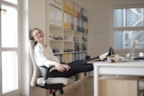 A woman wearing white and black work wear and red glasses reclines on an office chair in a bright office, her feet on the desk.