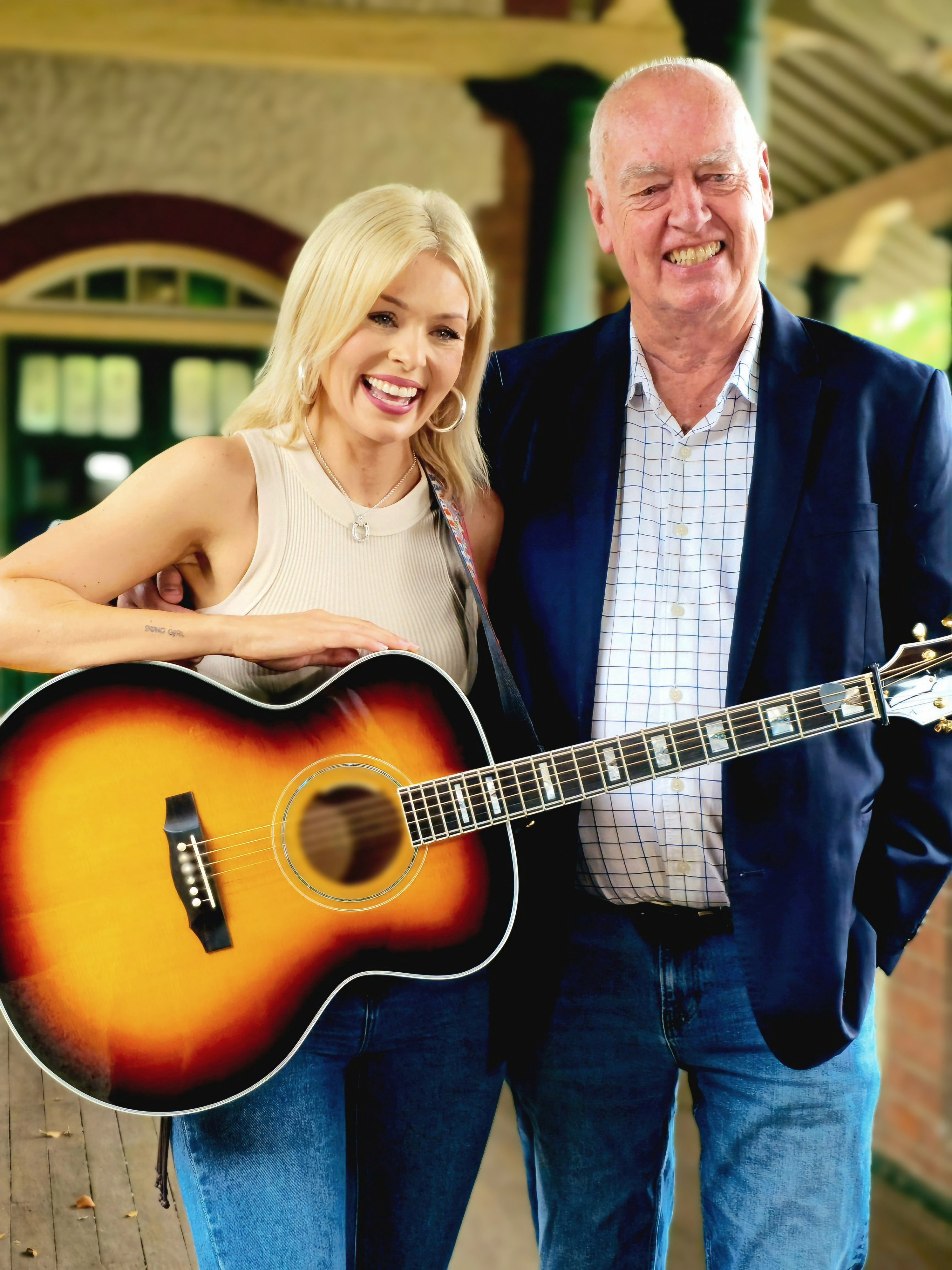 Uma mulher loira de camiseta e violão ao lado de um homem careca de jaqueta azul