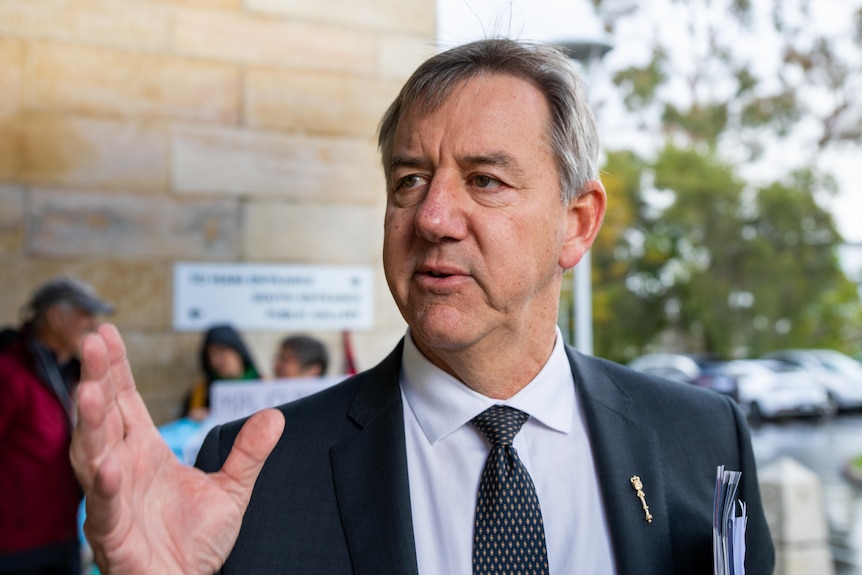 A man in a suit looking and gesturing to the left of frame.