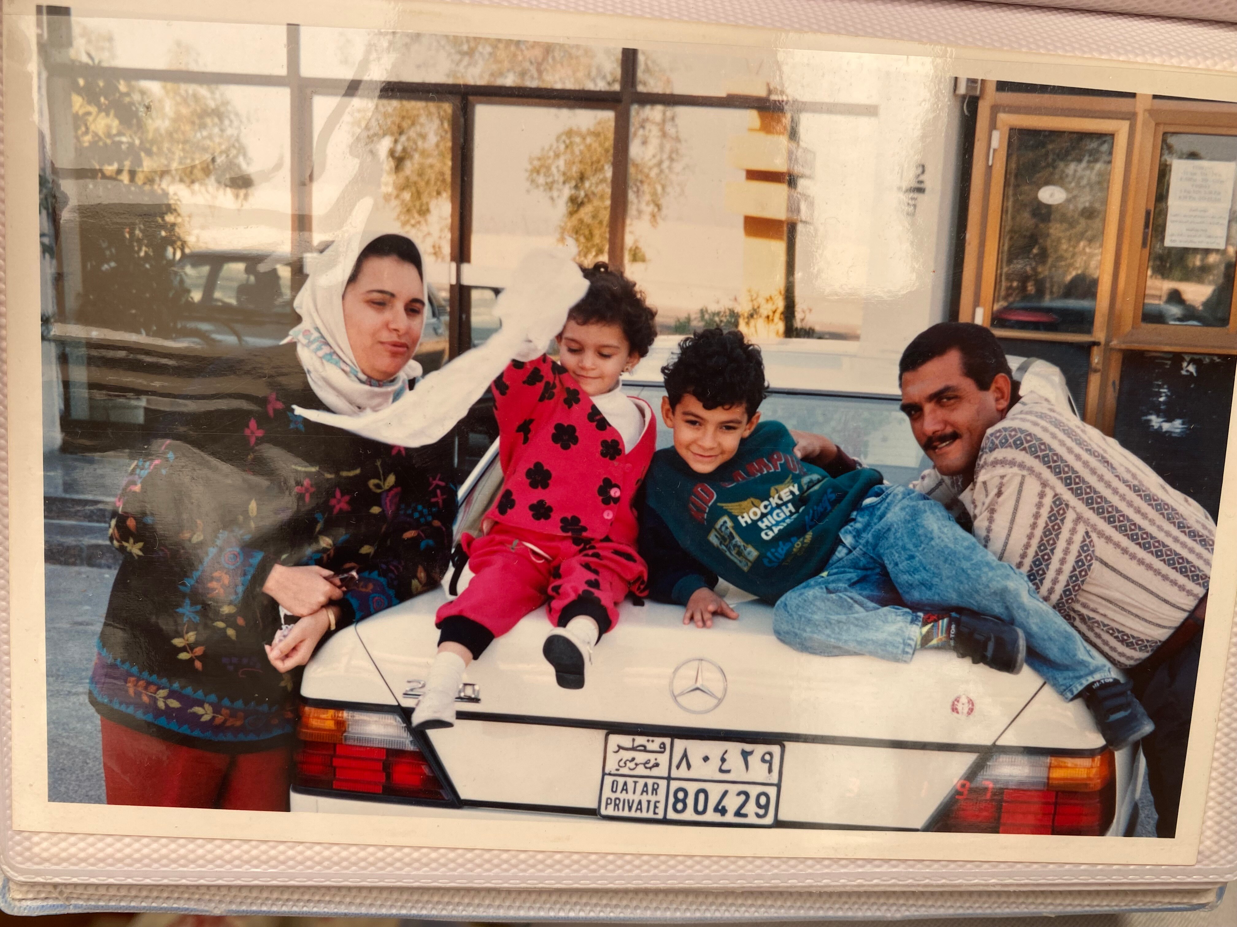 Two small children sitting on the boot of a white Mercedes with a man and a woman in a headscarf leading either side.