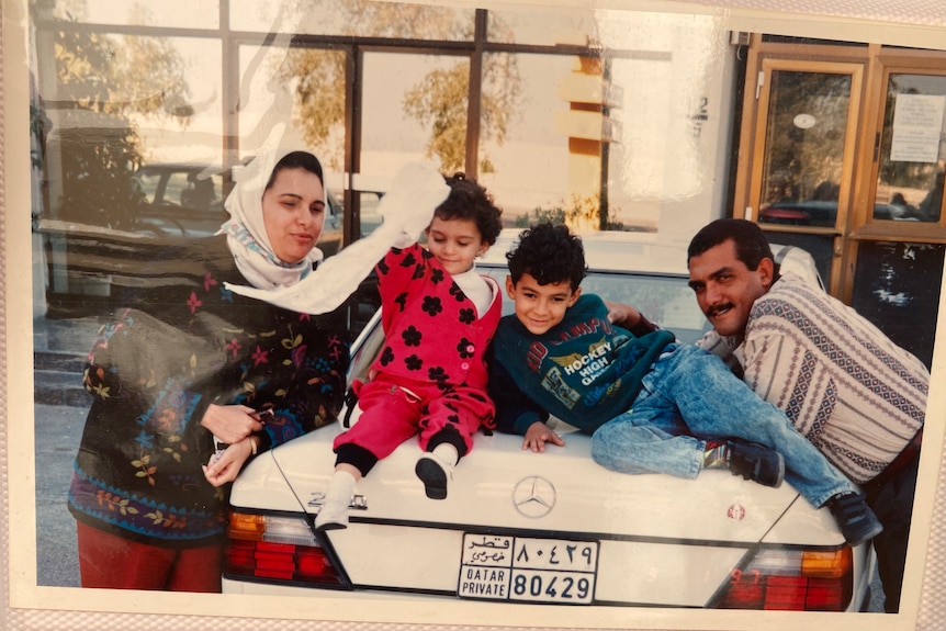 Two small children sitting on the boot of a white Mercedes with a man and a woman in a headscarf leading either side.