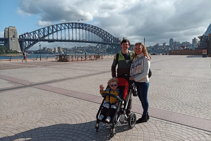 A woman and man with a child in a strollerpose for a photo in front of Sydney's Harbour Bridge.