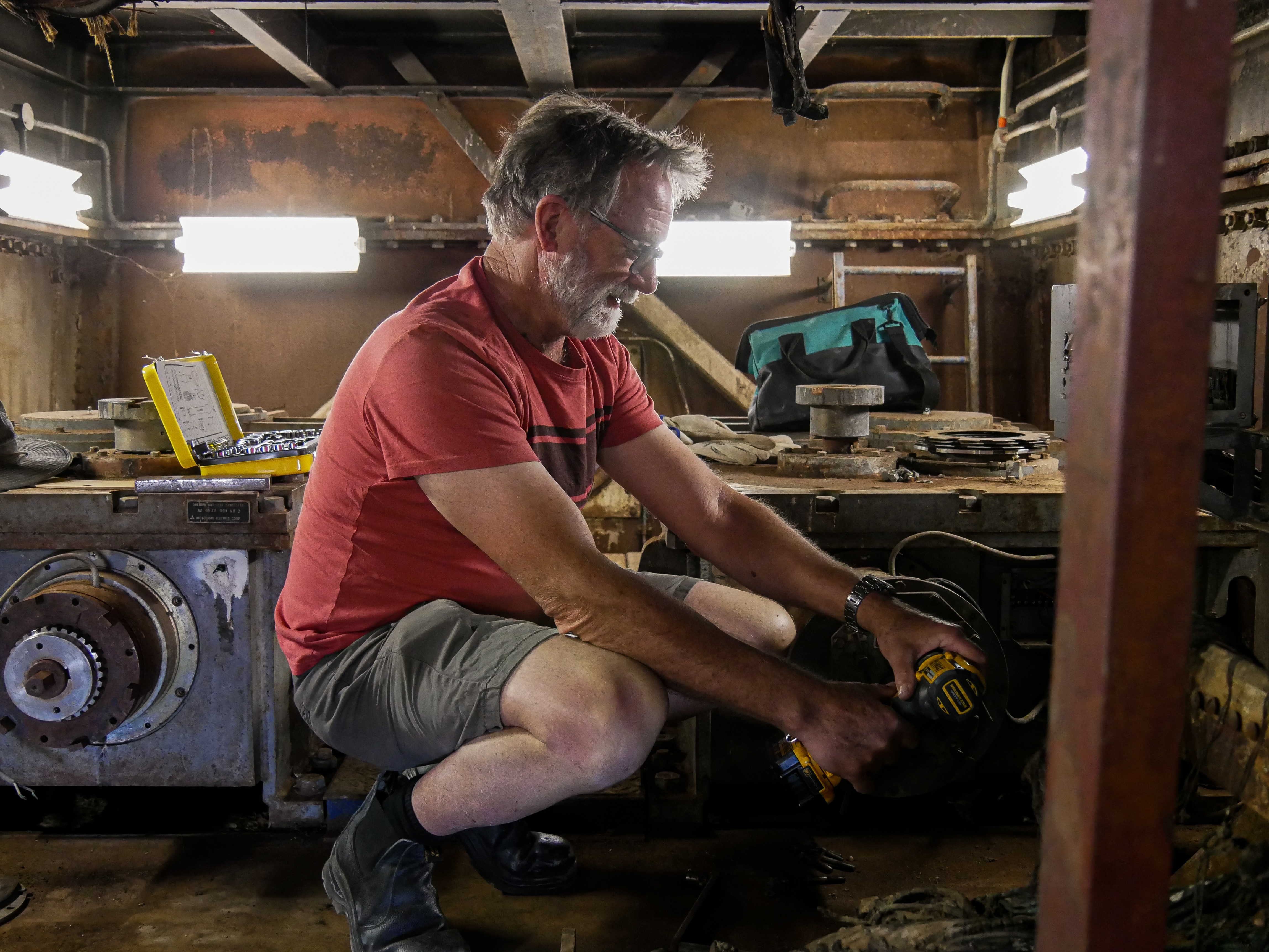 An older man with a beard holds a battery-powered drill to a machine.
