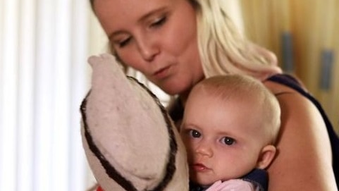 A blonde woman sits looking at her baby girl who is holding a doll