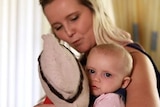 A blonde woman sits looking at her baby girl who is holding a doll