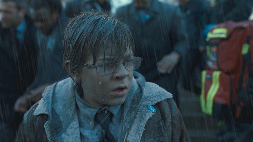 A young bespectacled boy looks upset and stands soaked in the rain near in front of crowd of onlookers in grey coats.
