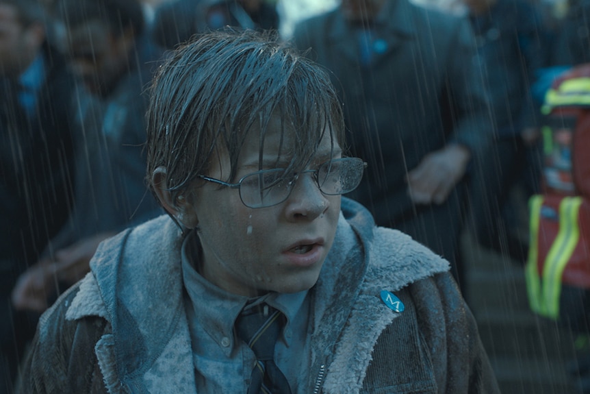 A young bespectacled boy looks upset and stands soaked in the rain near in front of crowd of onlookers in grey coats.