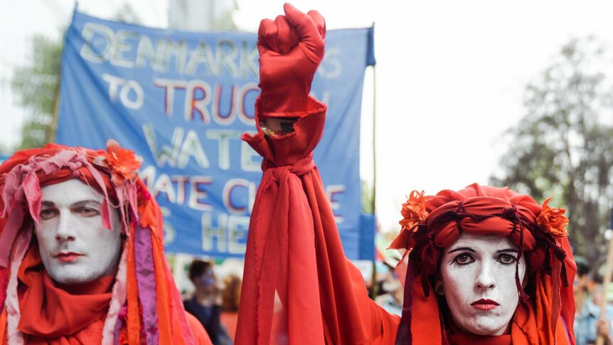Extinction Rebellion protesters dressed in red raise fists in front of a crowd waving banners.