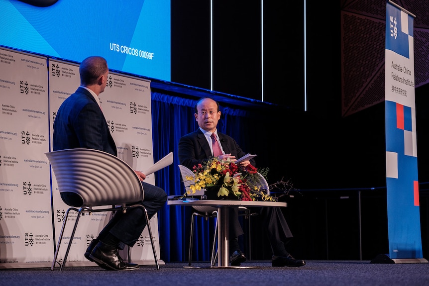 China's Ambassador to Australia Xiao Qian sitting on stage in front of flowers