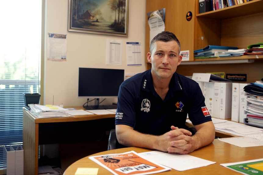 a man poses at a desk in a union t shirt, his fingers entwinned