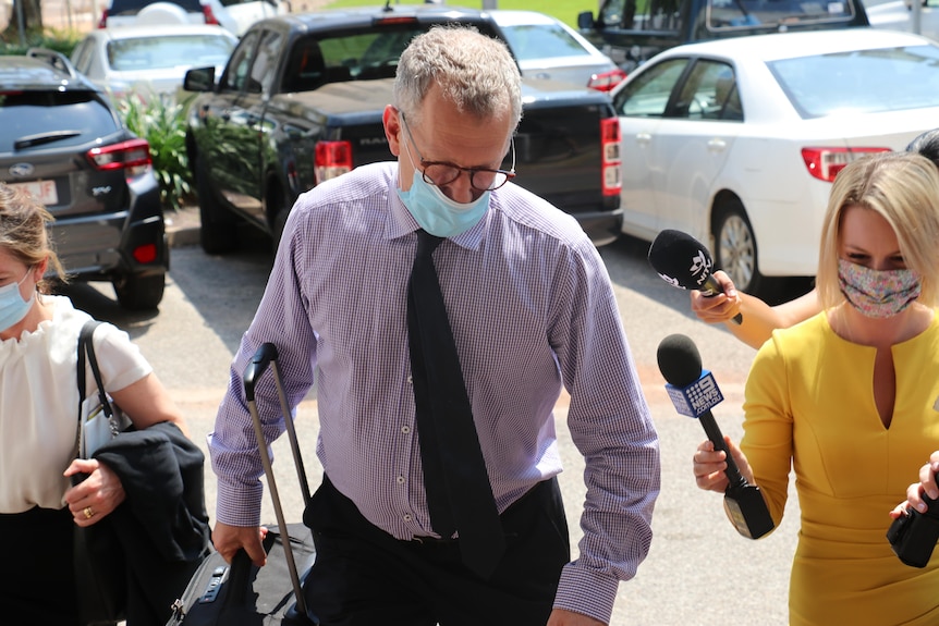 A man in a facemask and suit walks into court. Camera surround him.