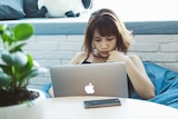 A young woman works on an Apple laptop in a white-bricked working space with indoor plants.