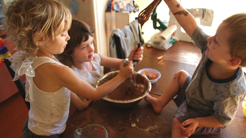 Two sisters mixing a bowl of chocolate cake batter and their messy toddler brother on the table, dribbling batter from whisk