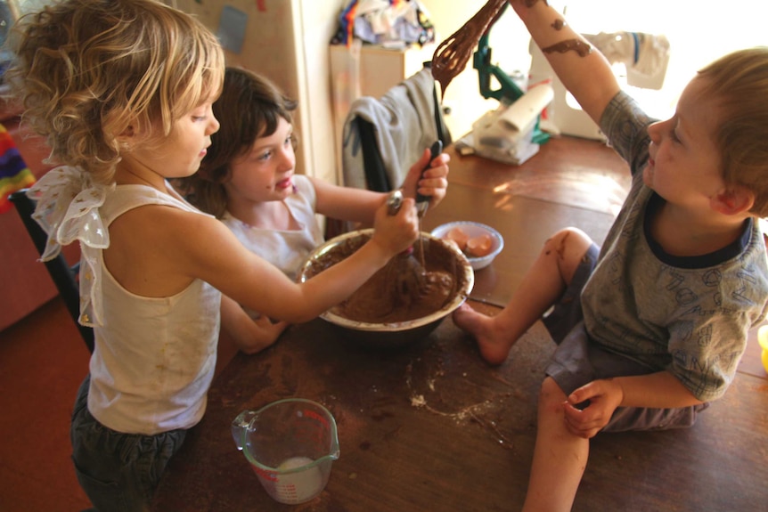 Two sisters mixing a bowl of chocolate cake batter and their messy toddler brother on the table, dribbling batter from whisk