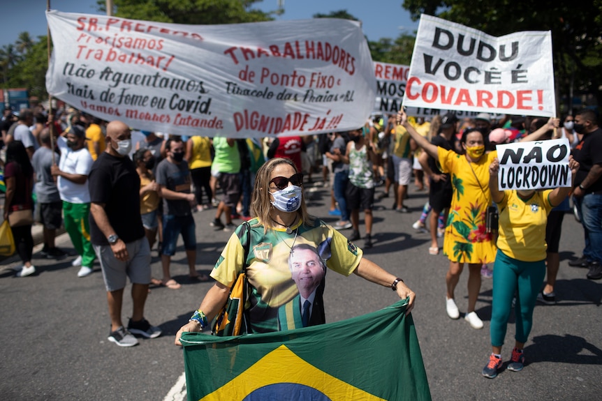 A middle-aged woman in face mask holds Brazilian flag at front of signs in street rally.